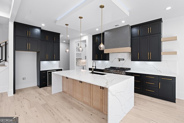 kitchen with pendant lighting, an island with sink, sink, light stone countertops, and wall chimney exhaust hood