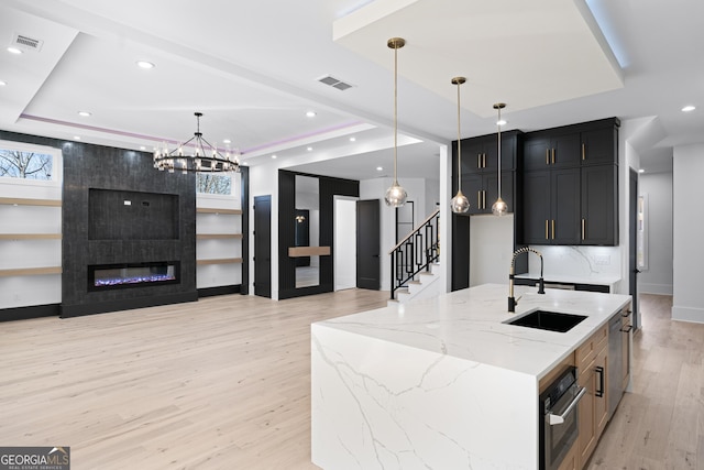 kitchen with sink, a center island with sink, a raised ceiling, light stone countertops, and oven