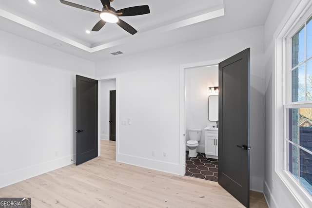 unfurnished bedroom with light hardwood / wood-style flooring, connected bathroom, ceiling fan, and a tray ceiling