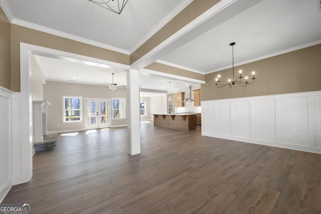 unfurnished living room featuring dark wood finished floors, wainscoting, ornamental molding, a decorative wall, and ceiling fan with notable chandelier