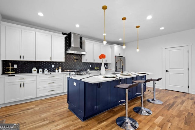 kitchen with wall chimney exhaust hood, hanging light fixtures, an island with sink, light hardwood / wood-style floors, and white cabinets