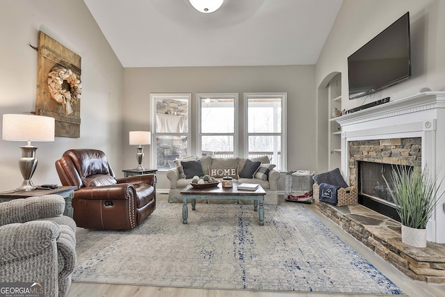 living room with built in shelves, lofted ceiling, a stone fireplace, and wood-type flooring