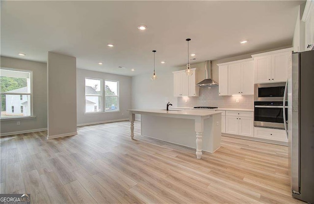 kitchen with a center island with sink, appliances with stainless steel finishes, pendant lighting, wall chimney range hood, and white cabinets