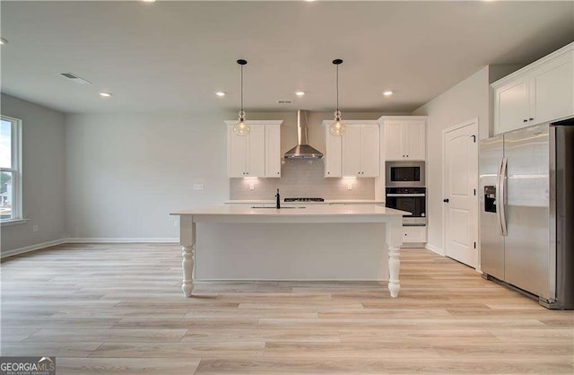 kitchen featuring wall chimney range hood, appliances with stainless steel finishes, white cabinetry, an island with sink, and decorative light fixtures