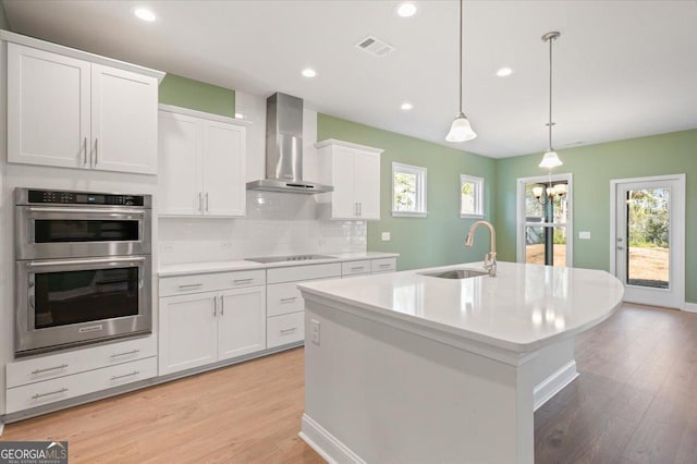 kitchen with sink, white cabinetry, hanging light fixtures, an island with sink, and wall chimney exhaust hood