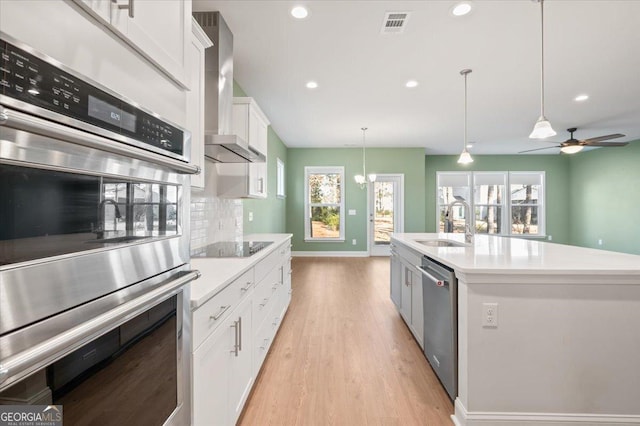 kitchen featuring white cabinetry, stainless steel appliances, a center island with sink, decorative light fixtures, and wall chimney exhaust hood