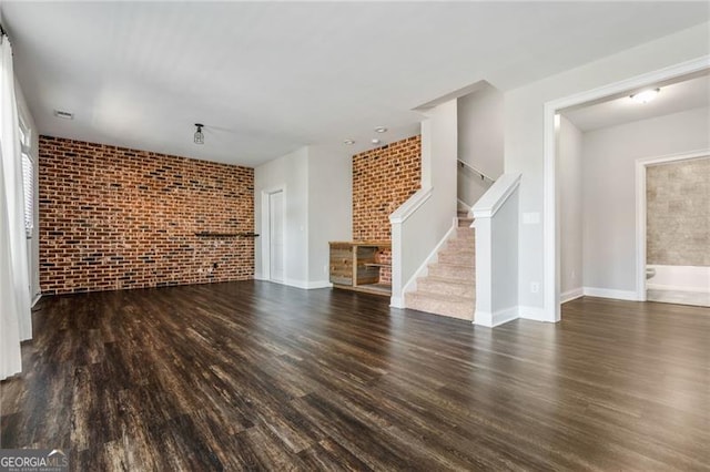 unfurnished living room featuring baseboards, dark wood finished floors, brick wall, and stairs