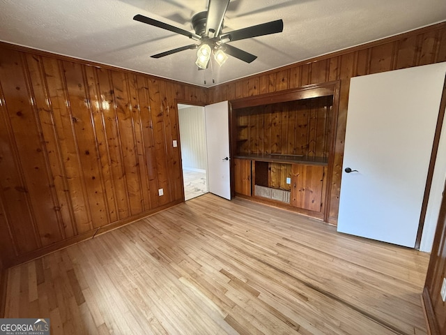 unfurnished bedroom with ceiling fan, a textured ceiling, light wood-type flooring, and wood walls