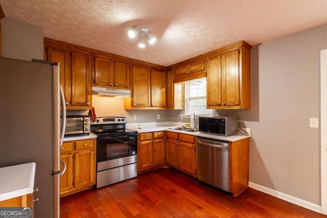 kitchen with dark wood-style floors, brown cabinets, stainless steel appliances, under cabinet range hood, and a sink