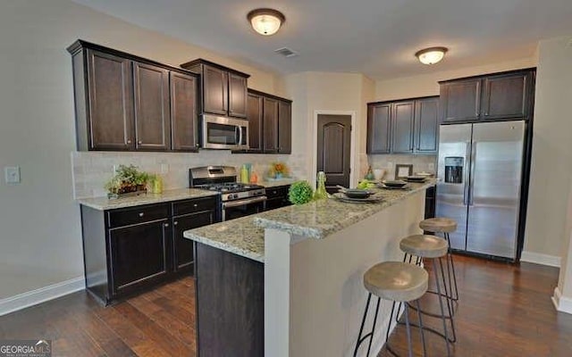 kitchen featuring dark brown cabinetry, a breakfast bar, light stone counters, stainless steel appliances, and backsplash