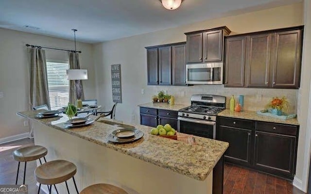 kitchen featuring a kitchen island with sink, hanging light fixtures, stainless steel appliances, dark brown cabinetry, and light stone countertops