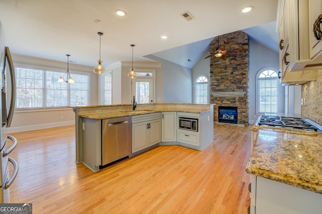 kitchen with a stone fireplace, a center island with sink, appliances with stainless steel finishes, pendant lighting, and light stone countertops