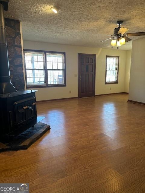unfurnished living room featuring a textured ceiling, baseboards, ceiling fan, wood finished floors, and a wood stove