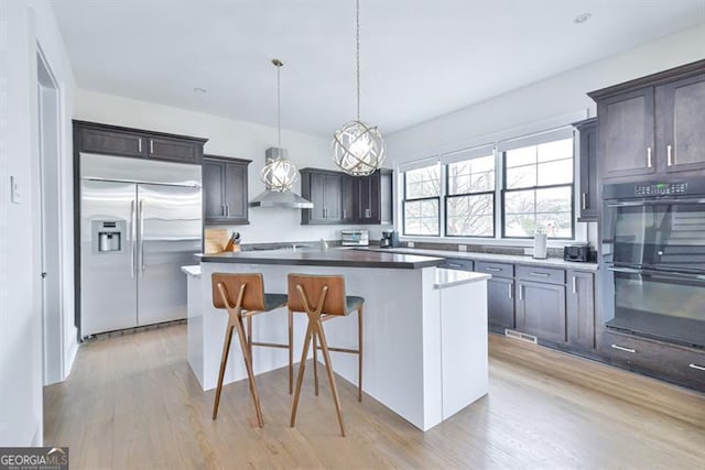 kitchen featuring built in fridge, decorative light fixtures, black double oven, a center island, and wall chimney range hood