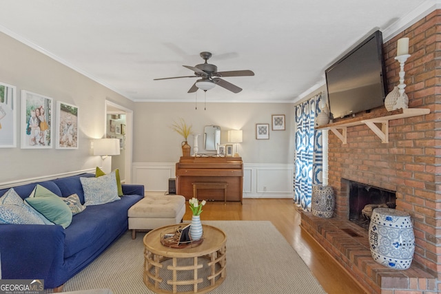 living area featuring crown molding, light wood-style flooring, a ceiling fan, a brick fireplace, and wainscoting