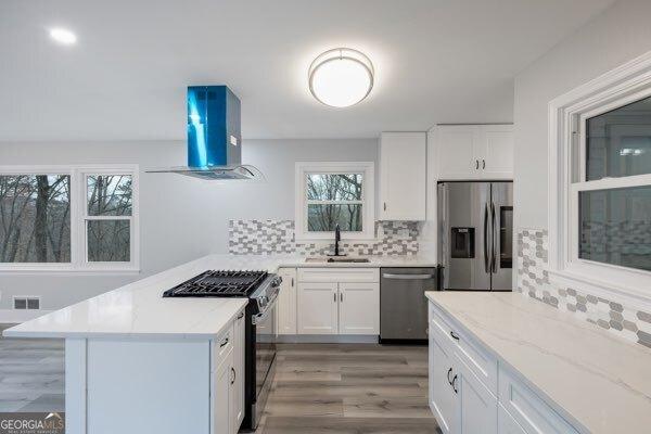 kitchen with sink, stainless steel appliances, light stone counters, island range hood, and white cabinets