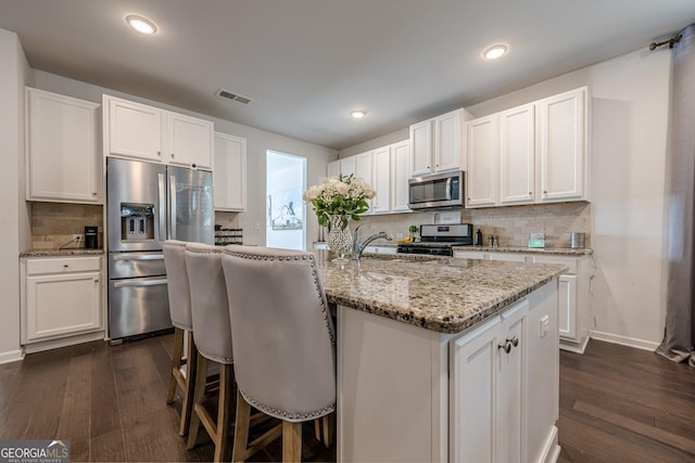 kitchen with dark wood-type flooring, light stone counters, stainless steel appliances, a kitchen island with sink, and white cabinets