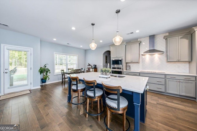 kitchen featuring appliances with stainless steel finishes, decorative light fixtures, tasteful backsplash, wall chimney range hood, and a center island with sink