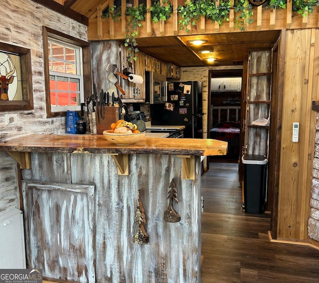 bar with dark wood-type flooring, wooden counters, black fridge, wooden ceiling, and wood walls