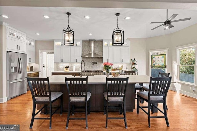 kitchen featuring white cabinetry, stainless steel fridge with ice dispenser, wall chimney range hood, and a large island with sink