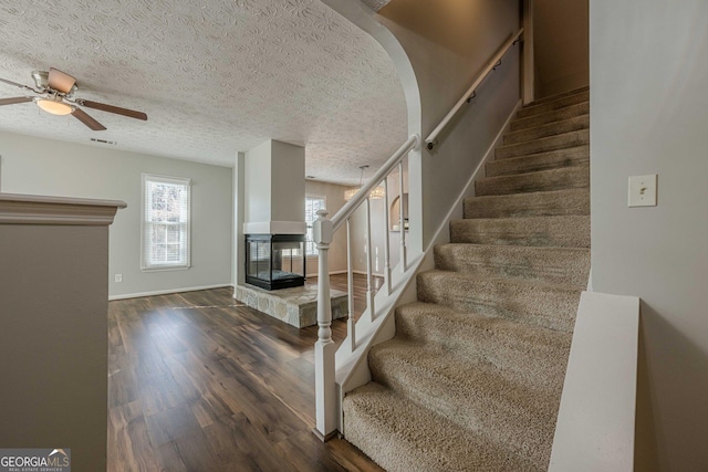 stairs with ceiling fan, wood-type flooring, a textured ceiling, and a multi sided fireplace