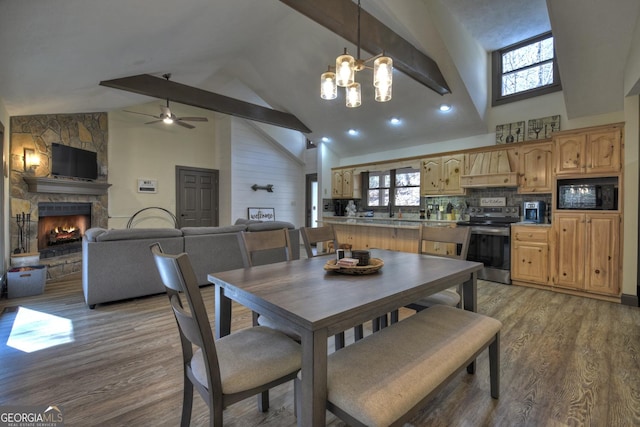 dining room with ceiling fan with notable chandelier, beam ceiling, high vaulted ceiling, light hardwood / wood-style floors, and a stone fireplace