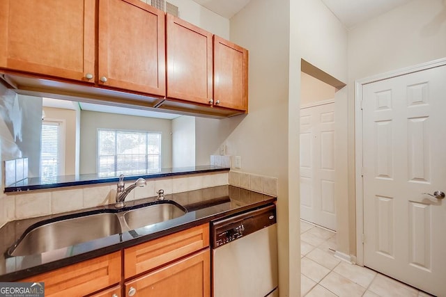 kitchen with brown cabinets, light tile patterned floors, dark countertops, stainless steel dishwasher, and a sink