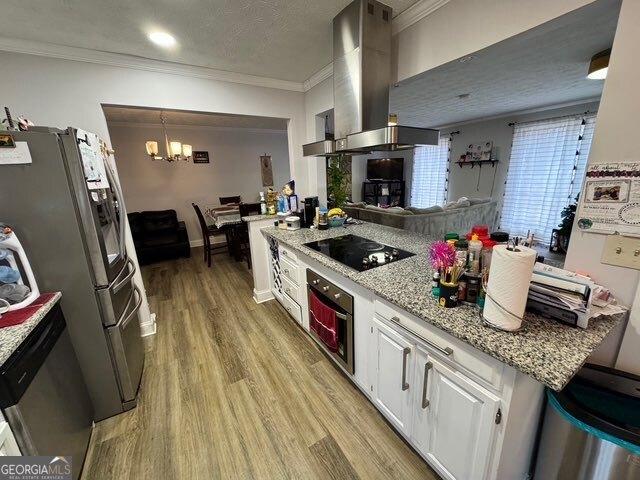 kitchen with appliances with stainless steel finishes, decorative light fixtures, white cabinets, wall chimney exhaust hood, and light hardwood / wood-style flooring