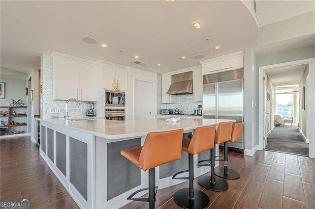 kitchen featuring dark wood-type flooring, wall chimney exhaust hood, sink, built in appliances, and a kitchen breakfast bar