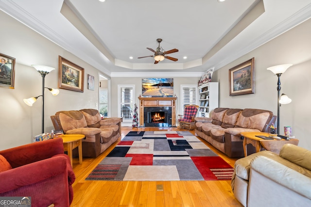 living room with ornamental molding, a raised ceiling, ceiling fan, a premium fireplace, and light hardwood / wood-style floors