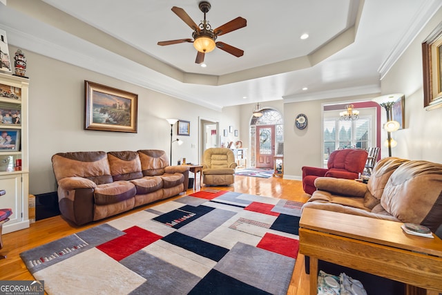 living room with crown molding, ceiling fan with notable chandelier, light wood-type flooring, and a tray ceiling