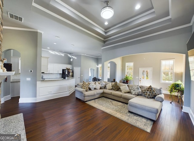 living room featuring a towering ceiling, dark wood-type flooring, ornamental molding, and a raised ceiling