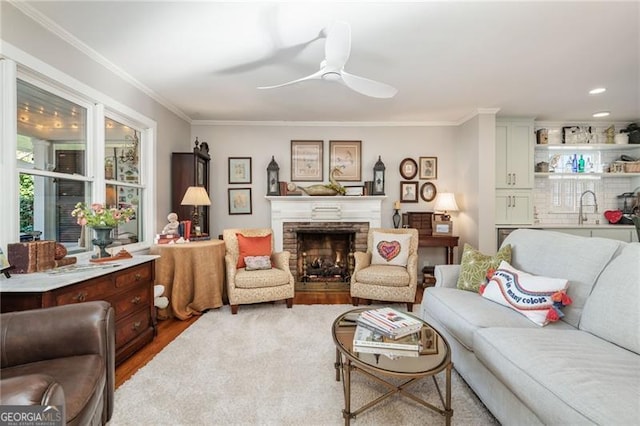 living area with a stone fireplace, wood finished floors, a ceiling fan, and crown molding