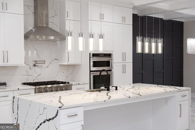 kitchen with wall chimney exhaust hood, coffered ceiling, decorative light fixtures, an island with sink, and white cabinets