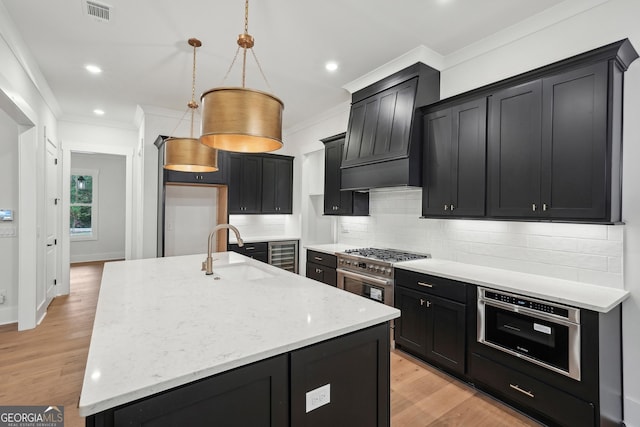 kitchen featuring premium range hood, a kitchen island with sink, backsplash, light stone countertops, and decorative light fixtures