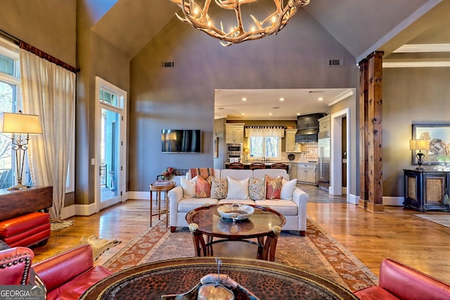 living room featuring high vaulted ceiling, decorative columns, a chandelier, ornamental molding, and light wood-type flooring