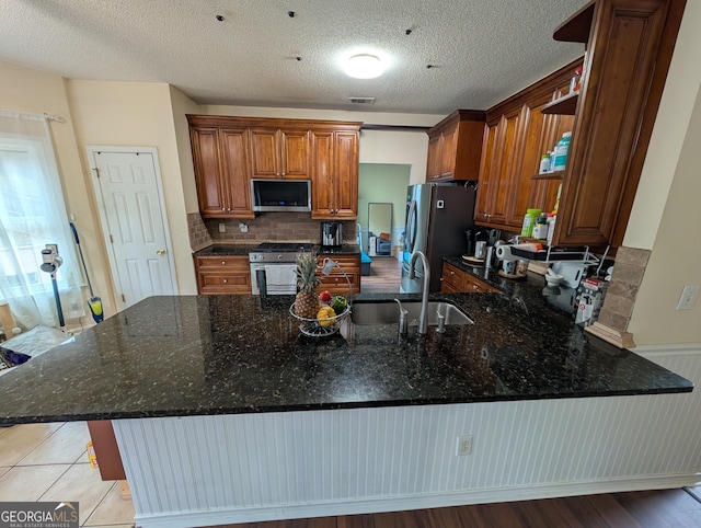 kitchen featuring a peninsula, a sink, appliances with stainless steel finishes, brown cabinets, and dark stone counters