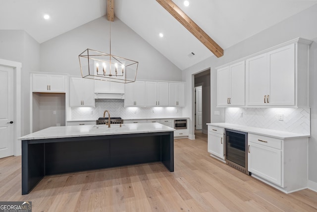 kitchen featuring white cabinetry, tasteful backsplash, a center island with sink, beamed ceiling, and beverage cooler