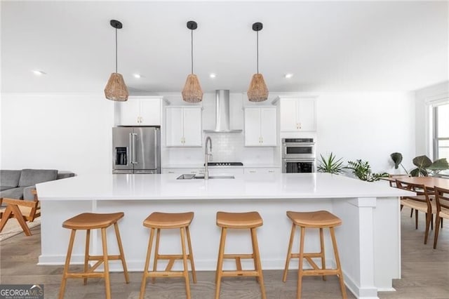 kitchen with white cabinetry, a large island with sink, stainless steel appliances, decorative backsplash, and wall chimney range hood