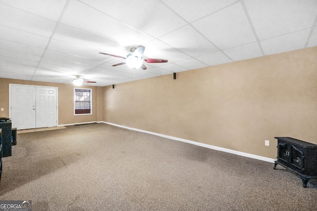 basement with ceiling fan, a drop ceiling, carpet, and a wood stove