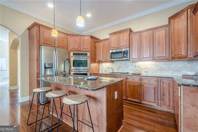 kitchen with stainless steel appliances, an island with sink, hanging light fixtures, and dark stone counters