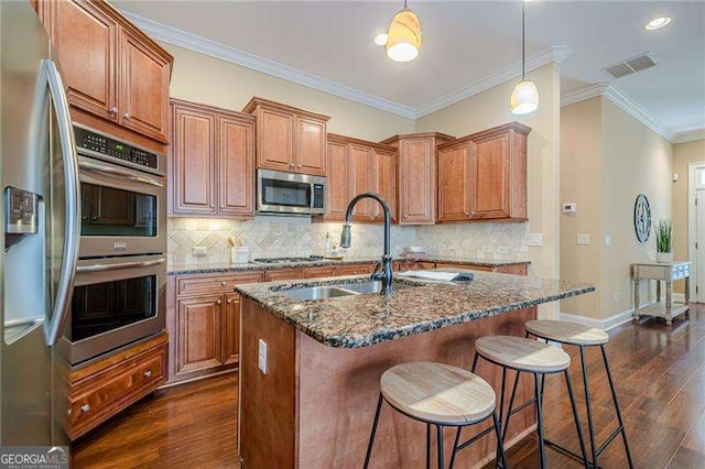 kitchen featuring sink, decorative light fixtures, appliances with stainless steel finishes, dark stone counters, and a kitchen island with sink