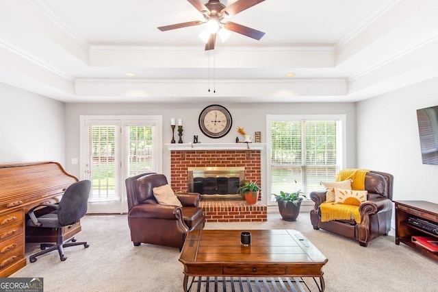 carpeted living room featuring crown molding, a brick fireplace, ceiling fan, and a tray ceiling