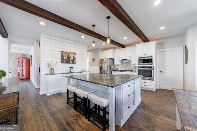 kitchen with pendant lighting, appliances with stainless steel finishes, white cabinets, dark stone countertops, and an island with sink