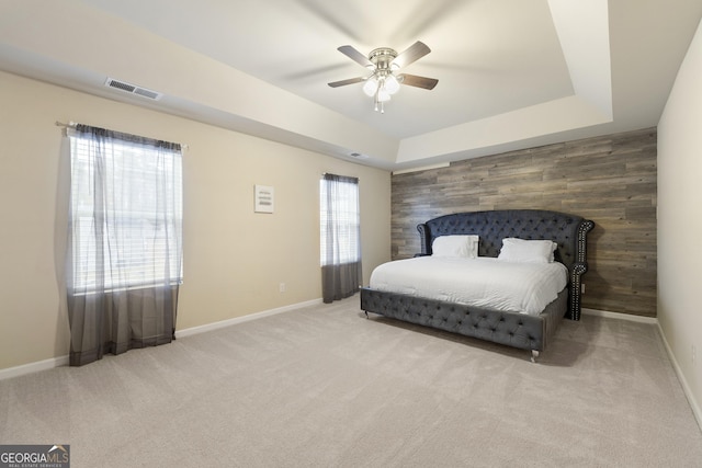 carpeted bedroom featuring multiple windows, a tray ceiling, wooden walls, and ceiling fan