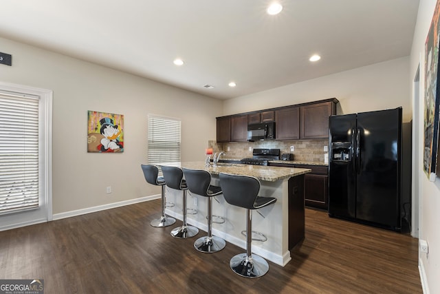 kitchen with dark wood-type flooring, a breakfast bar, dark brown cabinets, black appliances, and a center island with sink