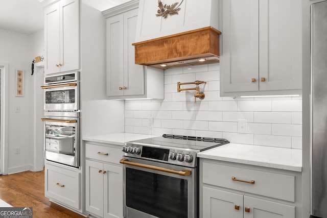 kitchen with premium range hood, appliances with stainless steel finishes, light stone countertops, hardwood / wood-style floors, and backsplash