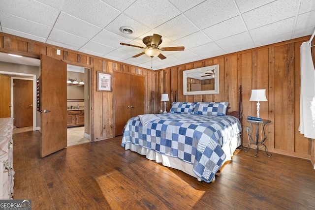 bedroom featuring ensuite bathroom, a paneled ceiling, wooden walls, hardwood / wood-style flooring, and ceiling fan