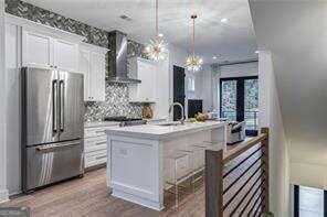 kitchen featuring wall chimney exhaust hood, hanging light fixtures, a center island with sink, appliances with stainless steel finishes, and white cabinets