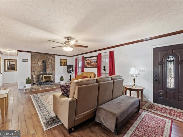 living room with wood-type flooring, a wood stove, ornamental molding, ceiling fan, and a textured ceiling